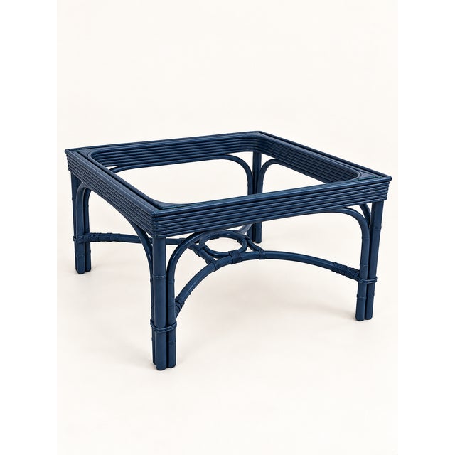 A sculptural rattan table presented in a deep navy finish, with curved rattan framework beneath a square glass top. Its...
