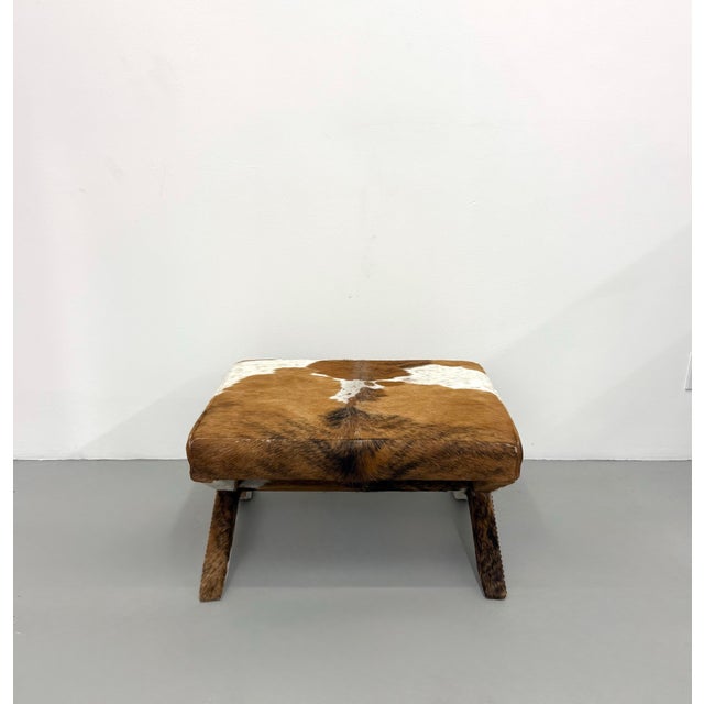 A striking vintage ottoman upholstered in genuine brown and white cowhide, featuring a clean square cushion atop an...