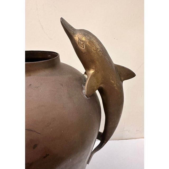 A sculptural brass vessel featuring two gracefully arched dolphin handles positioned symmetrically on either side of the...