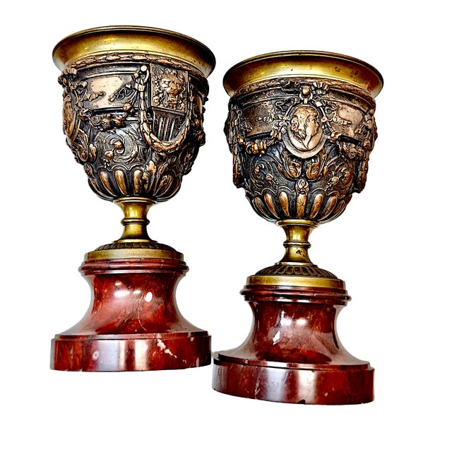 On offer is a pair of cast-bronze urns decorated with intricate reliefs, featuring brass rims and stems atop rouge marble...