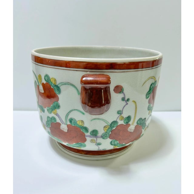 A vintage chinoiserie floral cachepot with double handles, soft green ground, rust-toned trim, and hand-painted botanical...