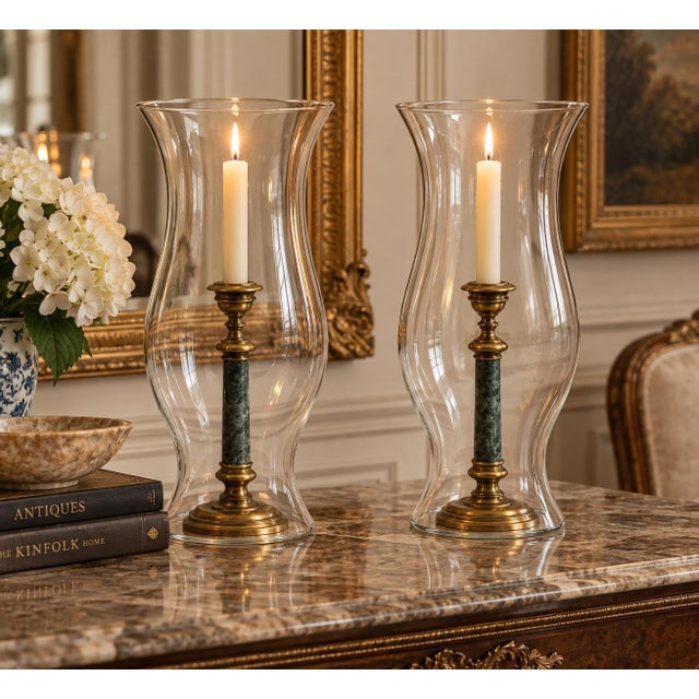 A matching pair of hurricane lamps featuring tall, hand-blown clear glass shades enclosing classical-style candlesticks....