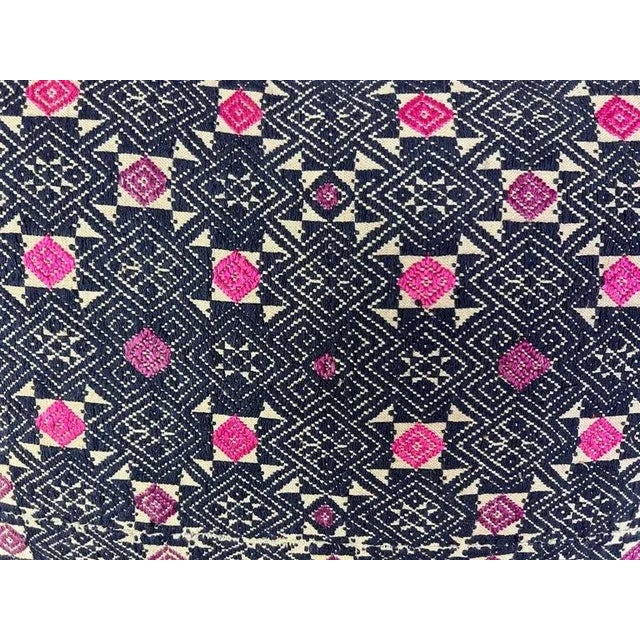 A decorative hand woven pillow with a geometric pattern. The primary colors in the pattern are navy & pink, with the pink...
