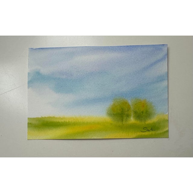 A serene landscape featuring rolling green fields beneath an expansive blue sky. Two softly blurred trees stand together...