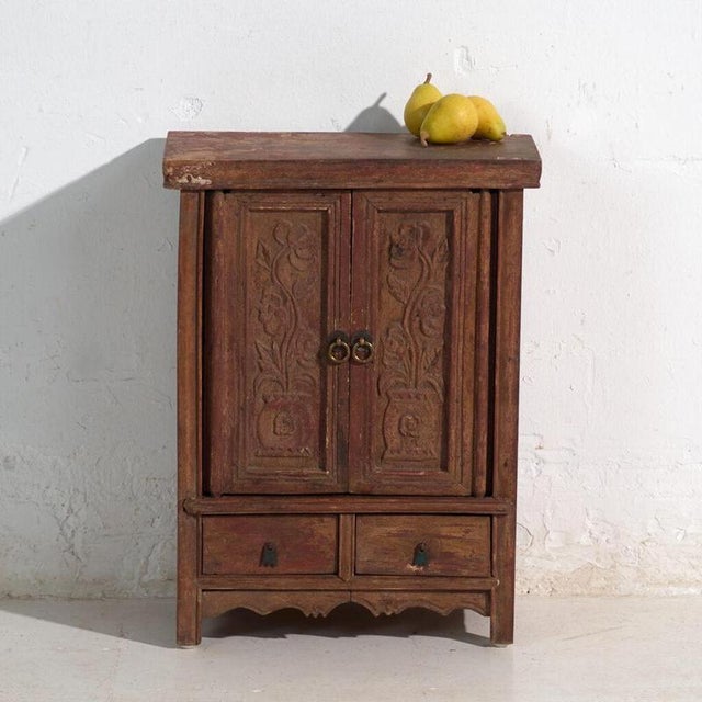 This antique Chinese cabinet in painted wood with two carved floral doors, two drawers and worn red lacquer, is a domestic...