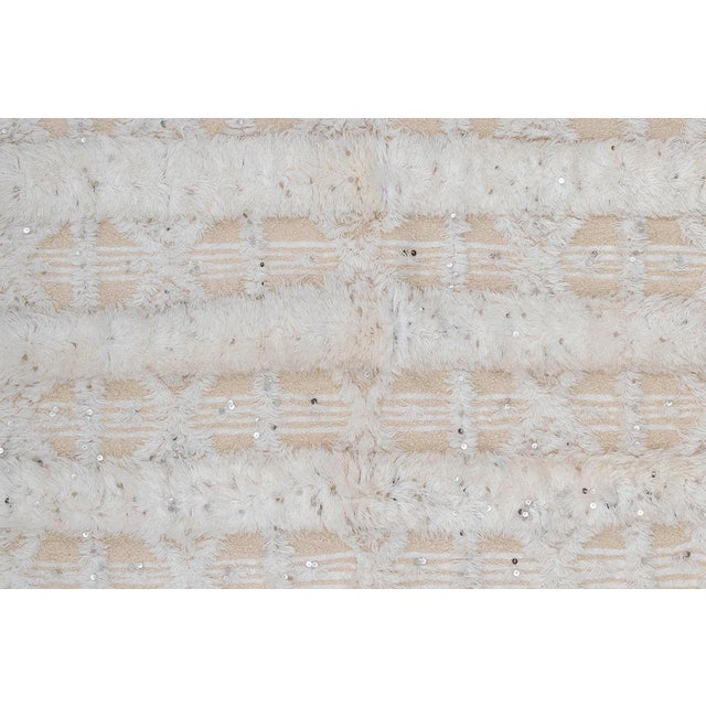 Artisanal hand-knotted rug features a cream-toned base with a raised textured pattern, highlighted by shimmering silver...