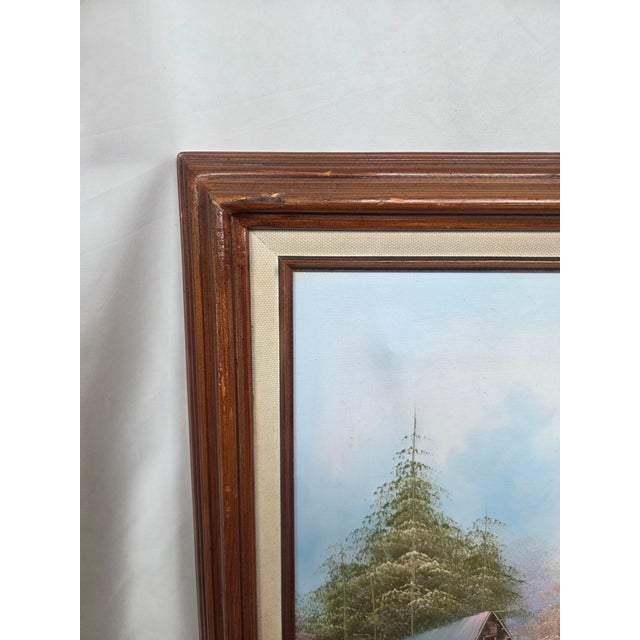 This vintage oil painting depicts a tranquil countryside landscape featuring a barn and cottage beside a calm lakeside...