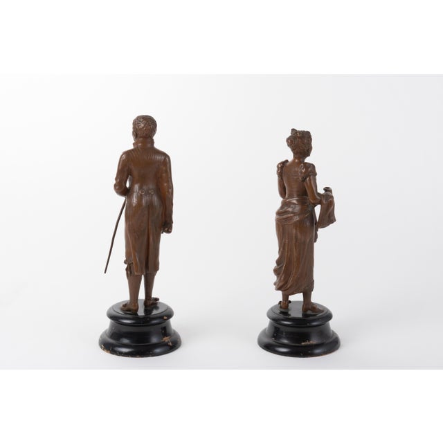 A pair of patinated bronze statues, depicting a man and a woman, both on dark, wooden, bases. The male figure, holding a...