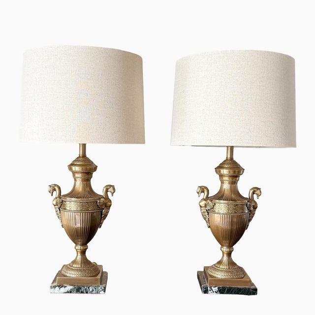 A striking pair of vintage brass table lamps in the Neoclassical style, featuring elegant swan-form handles and finely...