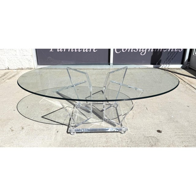 This contemporary glass-top table features a sleek, beveled circular surface resting on a striking sculptural acrylic...