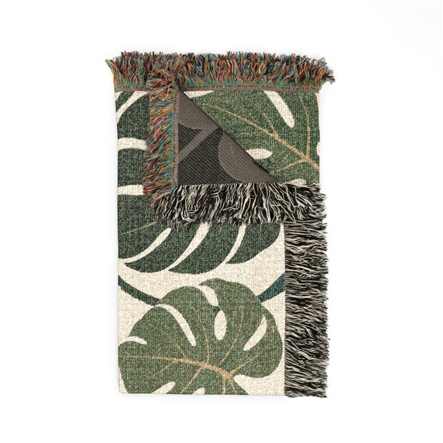 This monstera leaf woven throw blanket features oversized tropical foliage in deep teal and olive green with muted ochre...