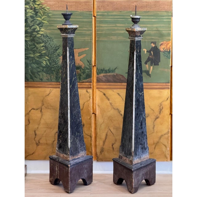 A pair of 19th century Continental floor prickets, each a tall tapering triangular column painted in black faux marble...