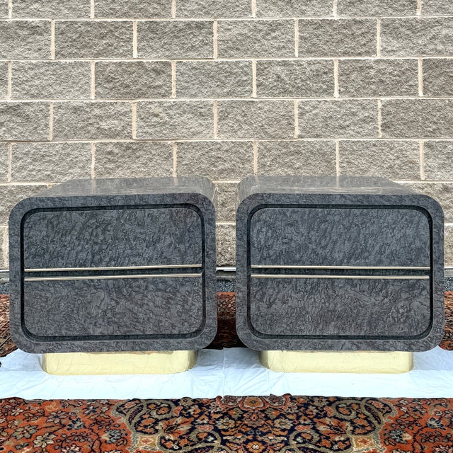 A pair of 1980s Postmodern 2 drawer nightstands in gray burl patterned laminate with brass trim and brass bases. They...