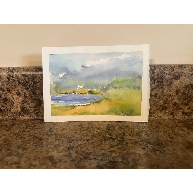 This gentle watercolor landscape depicts a quiet lakeside scene under a soft, cloud-streaked sky. The loose brushwork and...