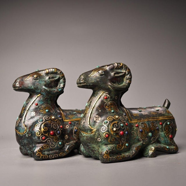 This is a matching pair of cast bronze vessels in the form of recumbent rams, featuring extensive gold and silver inlay...