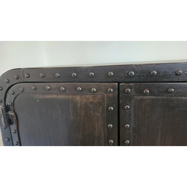 A striking industrial-style riveted metal sideboard featuring rounded corners, exposed rivet detailing, and a beautifully...