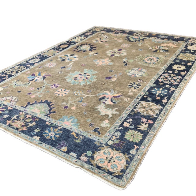 A refined modern Oushak rug featuring a warm tan field framed by a soft blue border, with large-scale floral motifs in...