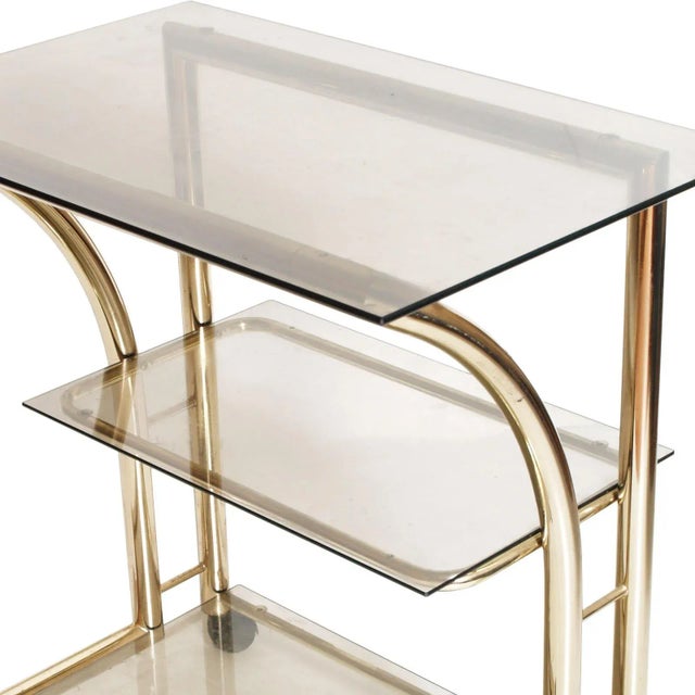 This Italian Mid-Century Modern 1970s bar cart or occasional table or console, in gilt brass, features 3 crystal 'fumè'...
