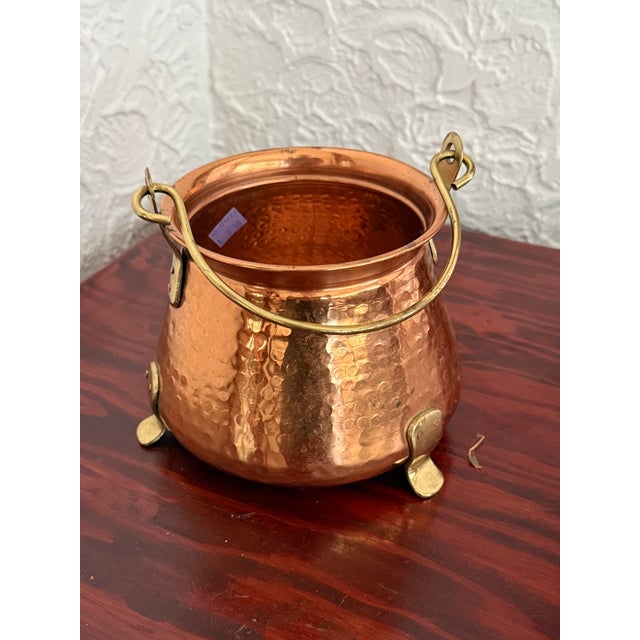 A small hand hammered copper cauldron with a brass handle and feet, warm and glowing on a hearth or in a window. Use it to...