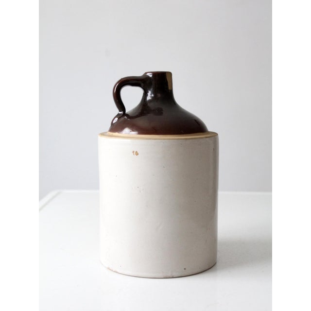 An antique American stoneware jug distinguished by a warm cream glaze with rich deep-brown glazing across the shoulder,...