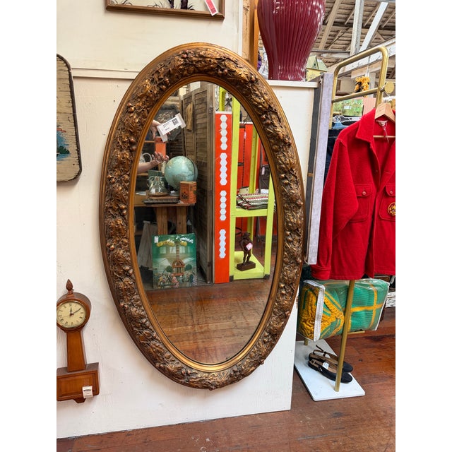Elegant oval wall mirror featuring an ornate raised floral and foliate frame with a richly aged gilt finish. Its intricate...