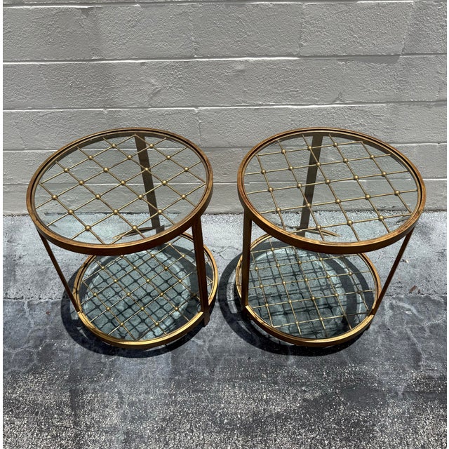 A refined pair of two-tier side tables featuring inset glass shelves framed by warm brass-tone metal and detailed with an...