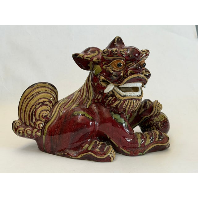 The glazed ceramic foo dog is portrayed crouching, with one paw resting and one on a ball. The back legs are relaxed and...