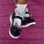 This artwork features a close-up view of a pair of black Mary Jane shoes adorned with decorative bows, resting on a...
