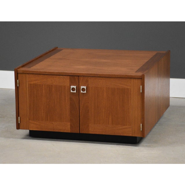 A mid century modern teak square coffee table with two doors, chrome finger pulls and an open bookshelf in the back side....