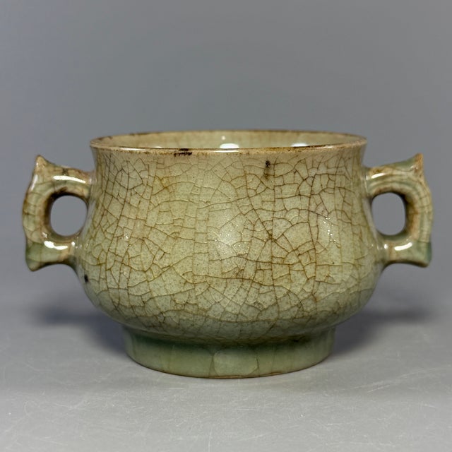 A vintage‑style Chinese Longquan celadon censer with two angular side‑handles. The whole surface is covered with natural...