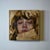 The painting "Bundled" captures a close-up of a figure with a contemplative expression, their face partially obscured by...
