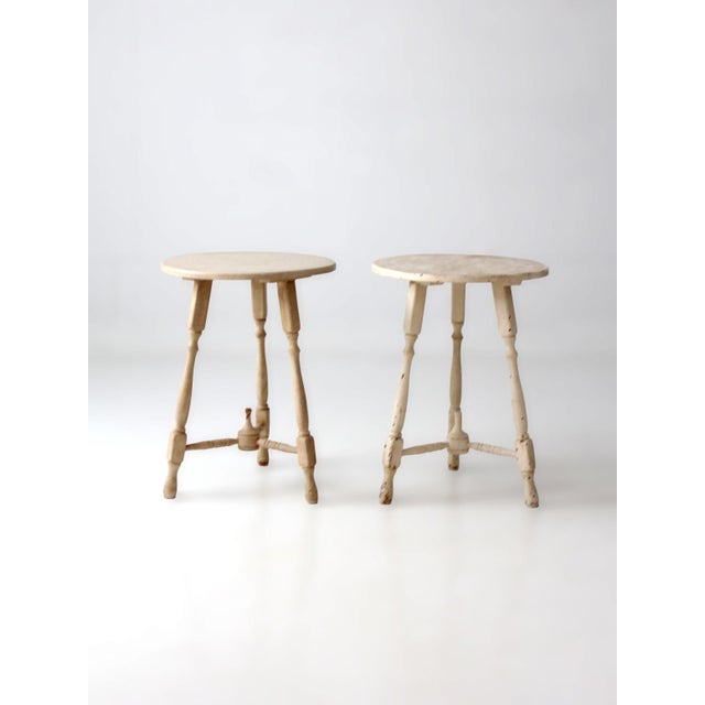 This pair of vintage pedestal side tables features softly distressed cream paint, round tops, and turned spindle legs...