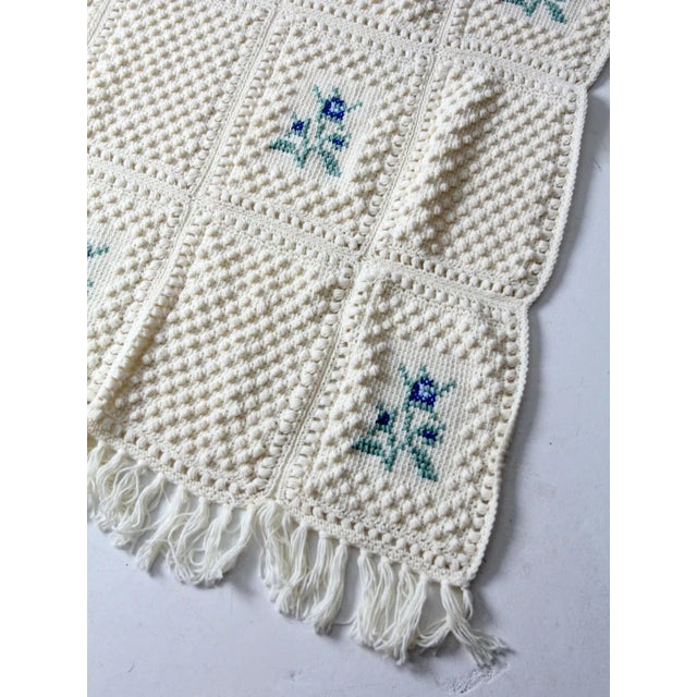 A vintage knit blanket in a soft cream tone, featuring bobble-knit square detailing and a central blue and green floral...