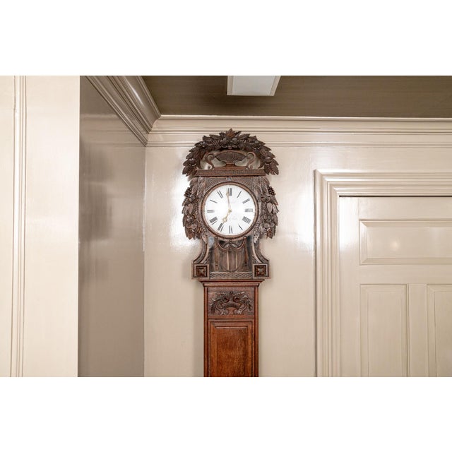 An antique French provincial tall oak longcase clock with warm oak tone, richly carved crest of foliage, flowers and a...