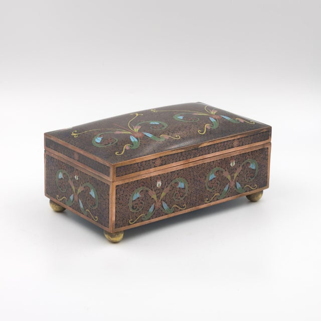 A rectangular cloisonné enamel box with gently domed lid, on four ball feet. The ground throughout is a dense dark brown-...