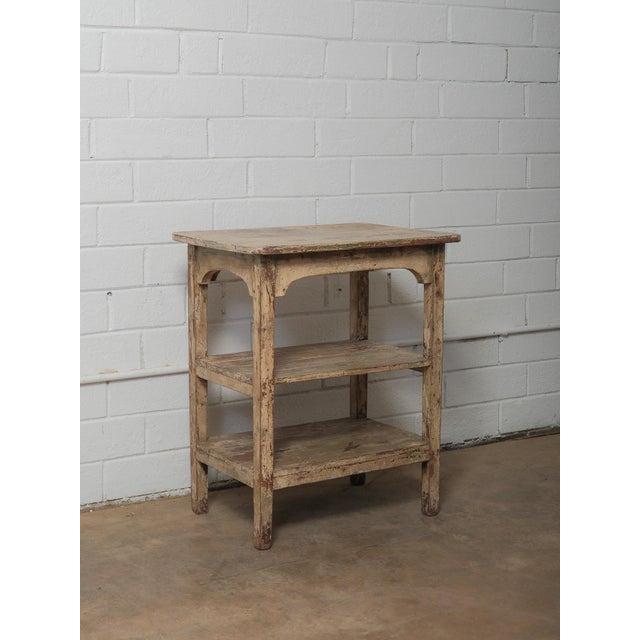A wooden table with two lower shelves, showing a layered surface built through long use and exposure. The rectangular top...