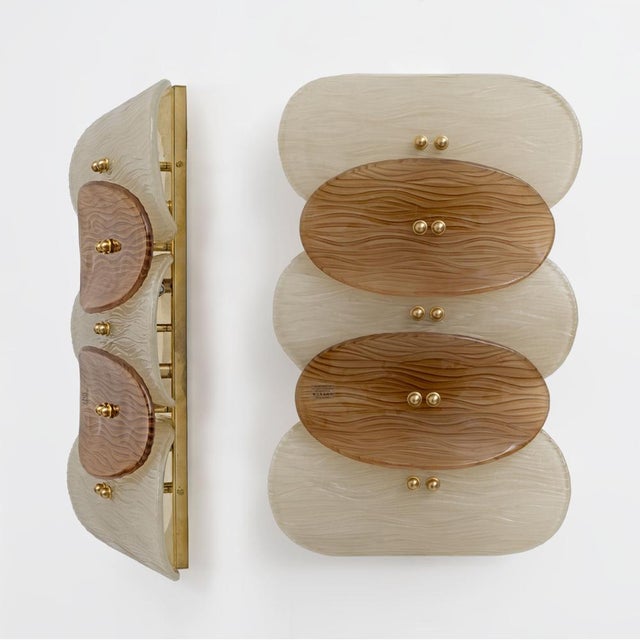 An elegant pair of wall lamps with polished brass frames and textured murano glass diffusers, featuring a refined wavy...