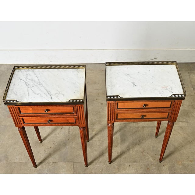 A pair of French, early to mid-20th century petite bedside tables offering two drawers in mahogany frames. Inset white...