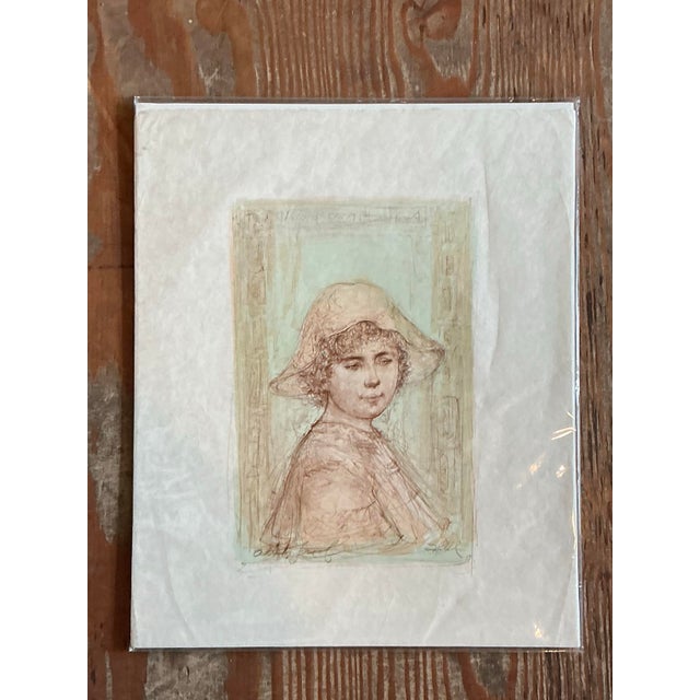 This vintage lithograph presents a quietly contemplative portrait of a young girl in a soft, wide-brimmed hat, her head...
