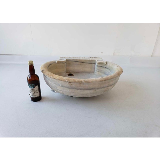 An antique, French carved marble wall mount basin, featuring a rounded bowl with a brass drain and brackets for mounting....