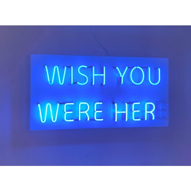 This artwork features a vibrant blue neon sign that boldly states "WISH YOU WERE HERE," creating an inviting yet...