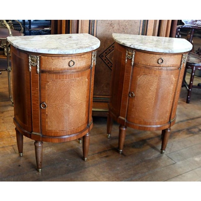 Elegant pair of Louis XVI style cabinets with demi lune forms and pale veined marble tops. Beautifully figured veneers are...