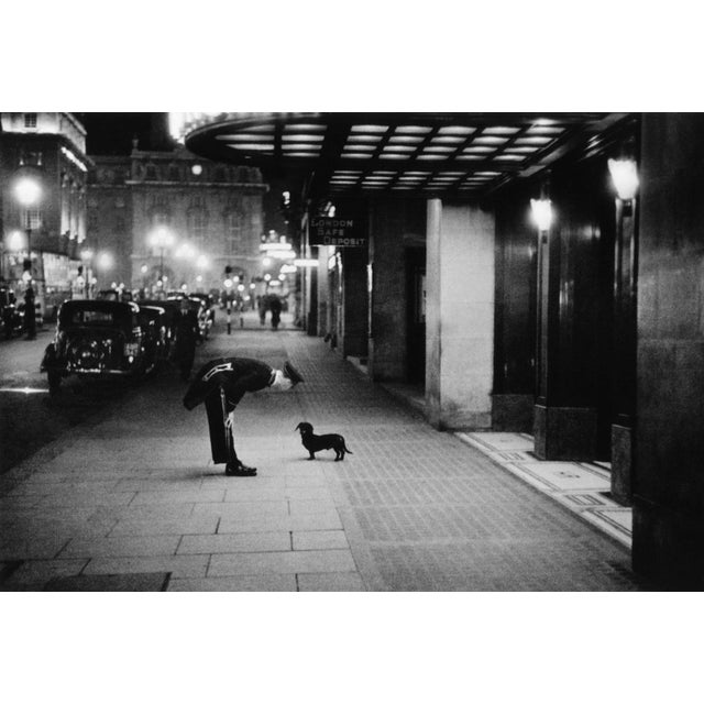 A hotel commissionaire talking to a small dachshund dog in Piccadilly Circus, London. Original Publication: Picture Post -...