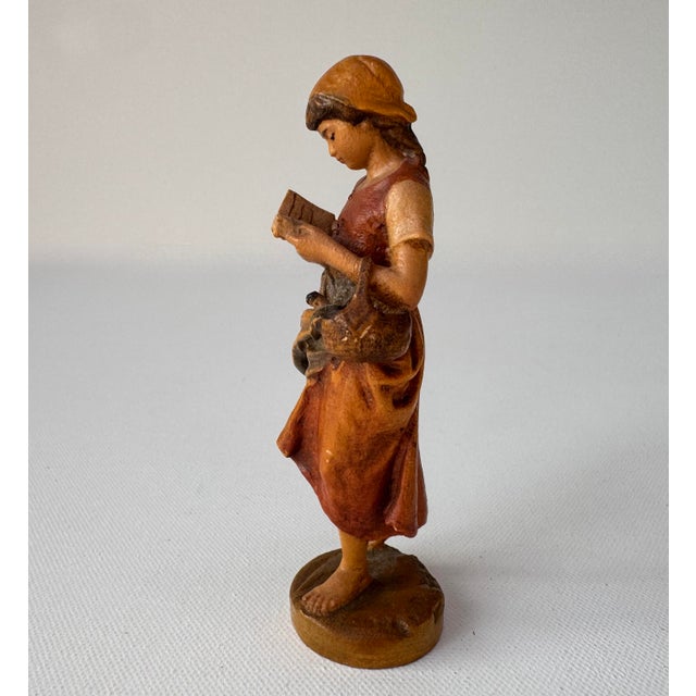 Detailed carved wooden figure of a barefoot girl absorbed in an open book, with a basket hanging from her arm. Her braided...