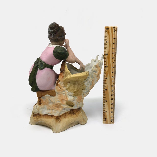 This elegant porcelain figurine features a Victorian-style woman in a graceful pink and green gown, leaning against a...
