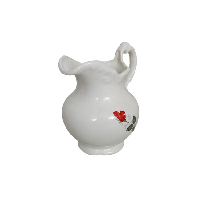 A white glazed ceramic pitcher, decorated on the left side with a single red rose on a slender stem with three leaves. The...