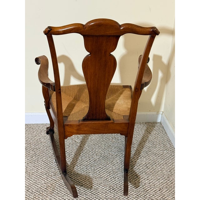 Antique wooden rocking chair featuring a Queen Anne style design with cabriole legs and a woven rush seat. Style: Queen...