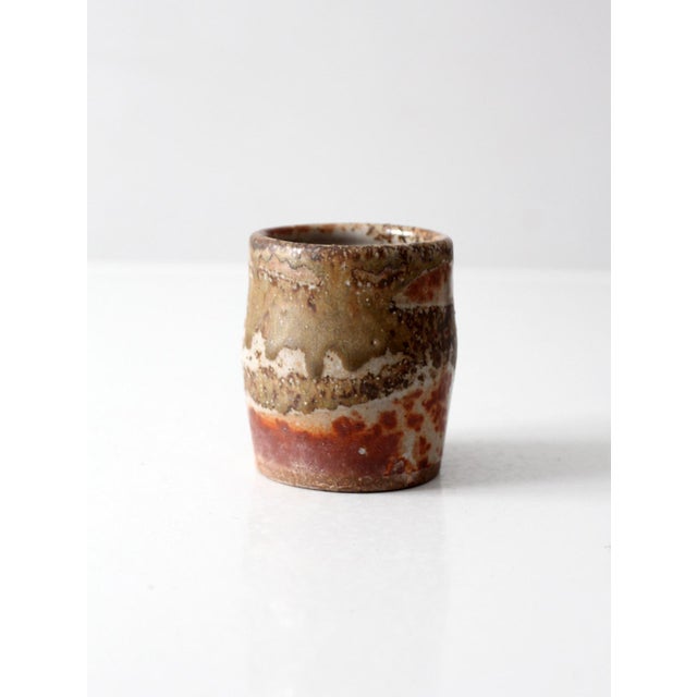 A vintage studio pottery cup with an organic, earth-toned glaze in layered brown, cream, and rust. The surface shows...
