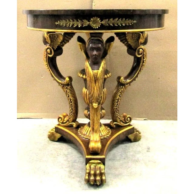 The Maitland Smith Egyptian Revival Table with Winged Creatures and Brass Claw Feet from the 1980s is a round accent table...