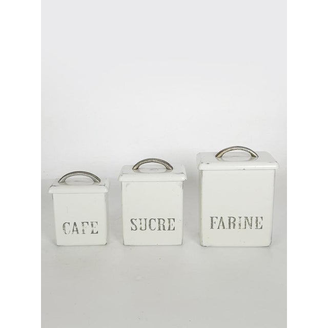 A charming set of three vintage kitchen canisters made of white-painted wood; each features a lid with a metal handle and...